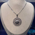 A silver pendant necklace featuring a detailed sun face design surrounded by small clear stones, displayed on a beige fabric jewelry bust against a dark blue background.
