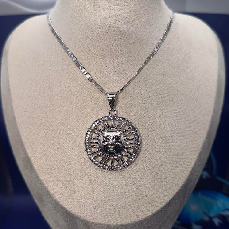 A silver pendant necklace featuring a detailed sun face design surrounded by small clear stones, displayed on a beige fabric jewelry bust against a dark blue background.