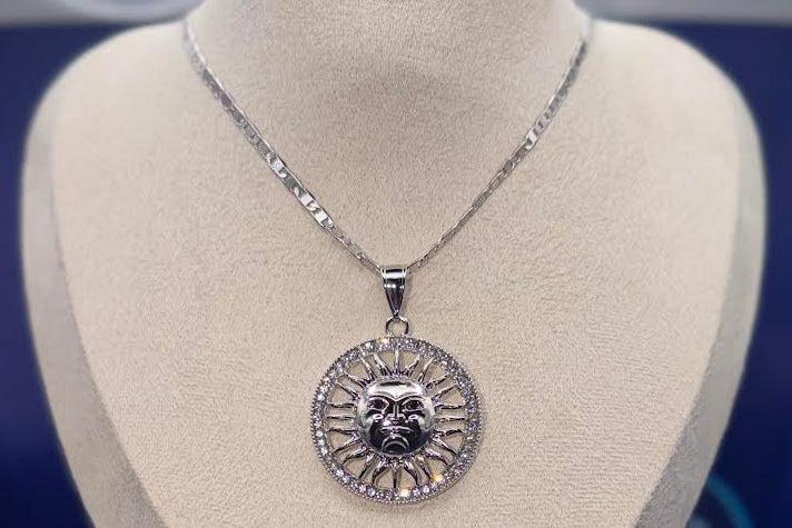 A silver pendant necklace featuring a sunburst design with a central dark emblem, displayed on a beige jewelry bust. The background is a deep blue, with a branded 'Azul Boutique' card visible in the lower left corner.